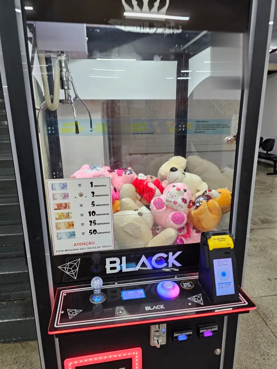 Detalhe da grua de pelúcias: vitrine com pelúcias, tabela de tentativas por valor, joystick e terminal de pagamento