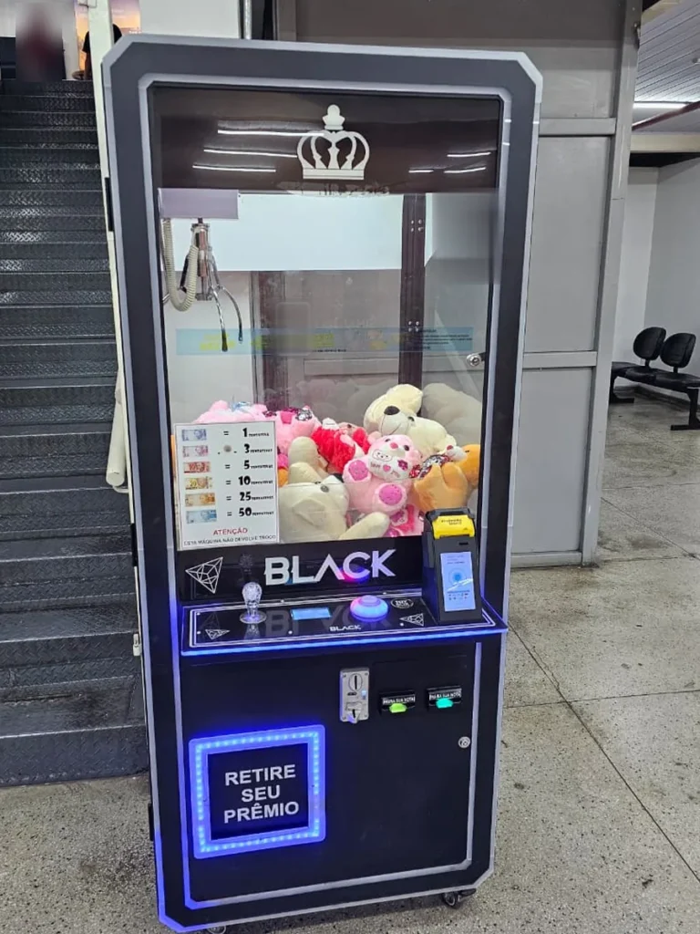 Máquina de grua de pelúcias PratikBox em área de circulação, com vitrine, joystick e retirada do prêmio